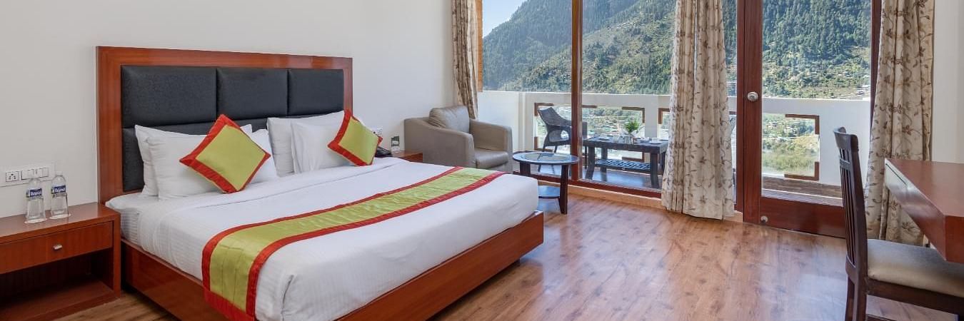 The image shows a hotel room with a large bed, a sitting area with a sofa and coffee table, and a large window with a view  | DLS Serenity Resort and Spa